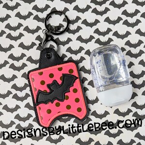 May include: A pink and black Halloween-themed hand sanitizer holder with a black bat design and a black keyring. The holder has a pink polka dot pattern. A clear hand sanitizer bottle with blue glitter is next to the holder. The text "designsbyLittleBee.com" is at the bottom.