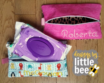 May include: Two zippered pouches with personalized names, "Roberta" and "McGowan", are shown on a wood surface. One pouch is pink with a leopard print lining and the other is blue with a white and blue animal print. A package of baby wipes is visible inside the blue pouch.