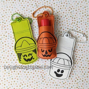 May include: Three Halloween-themed lip balm holders. One is lime green with a dripping face, one is orange with a jack-o'-lantern face, and one is white with a ghost face. Each has a matching keychain.