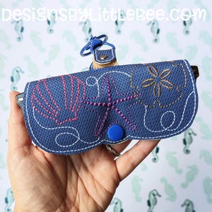 以下が含まれることがあります： 貝殻、ヒトデ、花の刺繍が施された青いメガネケース。ケースには青いスナップ留めとキーリングアタッチメントが付いています。「DesignsByLittleBee.com」の文字が上部に表示されています。