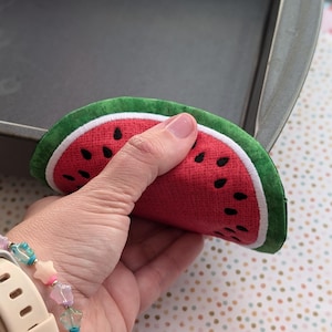 May include: A watermelon-shaped pot holder with a red center, black seeds, a white border, and a green rind. The pot holder is made of fabric and is designed to protect hands from heat.