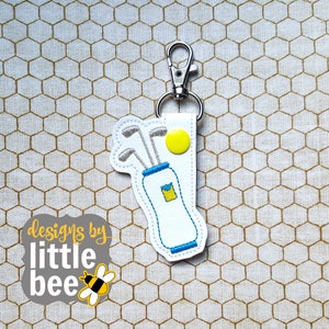 May include: White leather keychain with a blue golf bag and yellow golf ball. The keychain has a silver metal clip. The text "designs by little bee" is on the bottom of the keychain.
