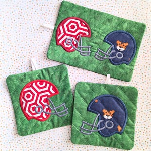 May include: Three green quilted pot holders with a football helmet design. Each pot holder features a different color helmet: red, blue, and navy blue. The helmets have a geometric pattern and a tiger face.