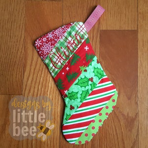 Peut inclure: Chaussette de Noël rouge et verte avec un motif patchwork. La chaussette est personnalisée avec le nom "Olivia" brodé en fil rouge.