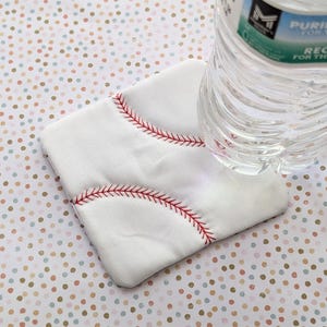 以下が含まれることがあります： 赤いステッチが施された、野球をテーマにした白いコースター。正方形のコースターは布製です。透明なプラスチック製のウォーターボトルがコースターの上に置かれており、水玉模様の表面に置かれています。