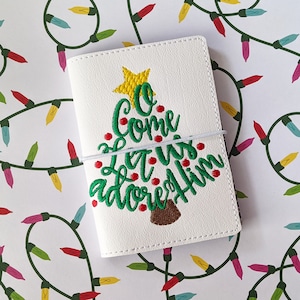 May include: White leather notebook cover with an embroidered green Christmas tree and the text "O Come Let Us Adore Him".