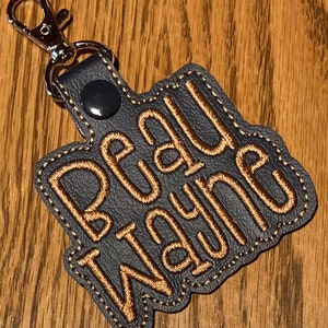 May include: A dark gray leather keychain with a silver metal clasp. The keychain has the name "Beau Wayne" embroidered in gold thread.