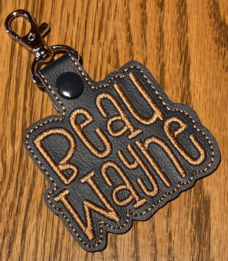 May include: A dark gray leather keychain with a silver metal clasp. The keychain has the name "Beau Wayne" embroidered in gold thread.