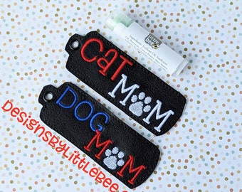 Puede incluir: Dos llaveros de cuero negro con costuras blancas. El llavero superior dice "CAT MOM" en letras rojas y blancas con una huella de pata blanca. El llavero inferior dice "DOG MOM" en letras azules y blancas con una huella de pata blanca. Los llaveros están sobre un fondo blanco con lunares marrones y verdes.