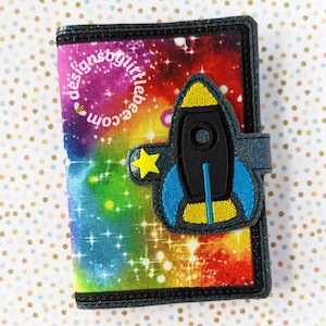 May include: A colorful wallet with a space theme. The wallet features a rocket ship applique with a yellow star and blue accents. The background is a vibrant mix of rainbow colors and stars, with the text "designsbylittlebee.com". The wallet has a black border.