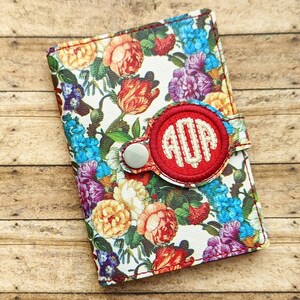 Op de afbeelding: Een portemonnee met bloemenprint met een rood monogramaccent. De portemonnee heeft een witte achtergrond met kleurrijke rozen, blauwe bloemen en groene bladeren. Het monogram staat in een rode cirkel met een knoopsluiting.