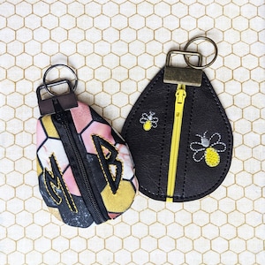 May include: Two small zippered pouches with key rings. One pouch is black leather with yellow embroidered bees. The other pouch is pink, black, and yellow fabric with the initials "MB" embroidered on it.