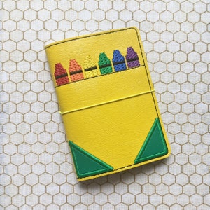 May include: A yellow leather notebook with a green triangle design and a row of embroidered crayons in rainbow colors.