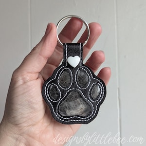 Op de afbeelding: Zwarte leren sleutelhanger met pootafdruk, witte stiksels en een zilveren sleutelring. Bovenop zit een witte hartvormige knoop. De pootafdruk bevat een lichtgekleurd materiaal. De tekst "designsbylittlebee.com" staat onderaan.