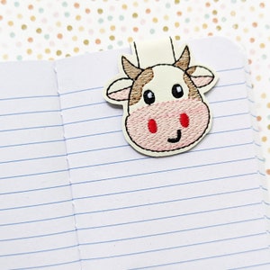 May include: A white and brown cow bookmark with red cheeks and black eyes. The bookmark is made of felt and has a magnetic closure.