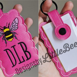 May include: A pink key chain with a bee and the initials DLB embroidered on it. The keychain has a clear plastic window for a card or photo.