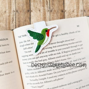 May include: A fabric bookmark featuring a green and red hummingbird design with a white border. The hummingbird is perched on a white tab, designed to slip over a page in a book. The bookmark is placed in an open book.