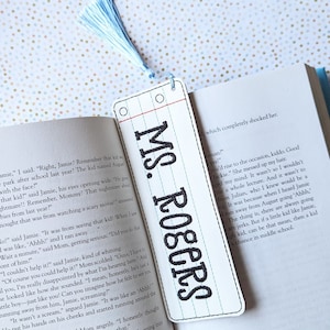May include: A white bookmark with black text that reads "MS. Rogers" and a blue tassel. The bookmark is designed to look like a school notebook page with red lines.