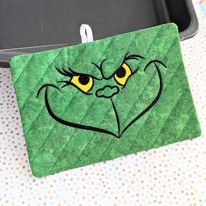 May include: Green quilted pot holder with an embroidered Grinch face. The Grinch has yellow eyes and a black mouth.