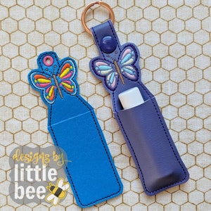 May include: Two blue and purple keychains with butterfly designs. The keychains are made of leather and have a pocket for a lip balm or other small item. The text "designs by little bee" is visible in the bottom left corner of the image.