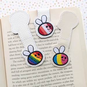 Puede incluir: Tres marcapáginas de cuero blanco con diseños de abejas bordadas. Las abejas son coloridas y tienen diferentes patrones. Una abeja es arcoíris, otra es roja, blanca y rosa, y otra es azul, rosa y amarilla.