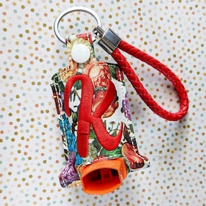 May include: A floral print keychain holder with a red braided wrist strap. The holder features a large embroidered red letter "R" and a white snap closure. An orange object is visible at the bottom.