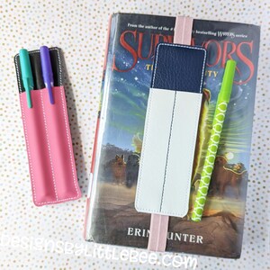May include: A pink pencil holder with two pens inside, and a white and blue bookmark with a pink elastic band attached to a book titled "Survivors" by Erin Hunter.