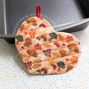 May include: A heart-shaped quilted pot holder with a fall-themed fabric featuring mushrooms, leaves, and stripes. The pot holder has a red loop for hanging.