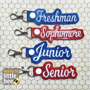 May include: Four keychains with glitter fabric and white text. The keychains are labeled "Freshman", "Sophomore", "Junior", and "Senior".