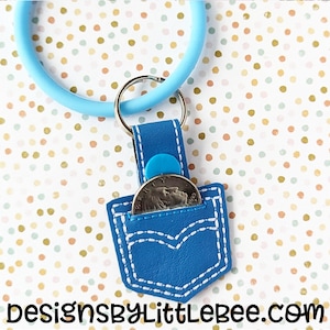 May include: A blue leather keychain with a small pocket that can hold a coin. The pocket has white stitching and a white stitched design that resembles a pair of jeans. The keychain has a blue ring and a silver key ring.