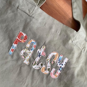 May include: A sage green tote bag with the name "Patsy" appliquéd in colourful floral fabric. The bag has long handles and is made of a sturdy canvas material, ideal for carrying items.