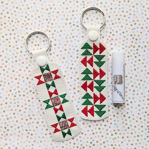 Puede incluir: Dos llaveros de cuero blanco con patrones geométricos rojos y verdes. El llavero de la izquierda tiene un patrón de estrella y el llavero de la derecha tiene un patrón de triángulo. Un pequeño tubo de bálsamo labial de menta está en el fondo.