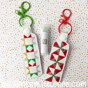 Puede incluir: Dos llaveros de tela blanca con diferentes diseños bordados. Un llavero tiene un patrón geométrico rojo, verde, dorado y plateado. El otro llavero tiene un patrón de molino de viento rojo y verde. Ambos llaveros tienen un botón blanco y un anillo de metal.