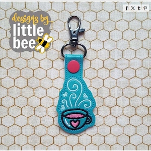 Könnte beinhalten: Ein türkisfarbener Schlüsselanhänger mit einem silbernen Verschluss und einem rosa Knopf. Der Schlüsselanhänger zeigt ein gesticktes Design einer Kaffeetasse mit Herz und Dampf sowie den Text "designs by little bee".