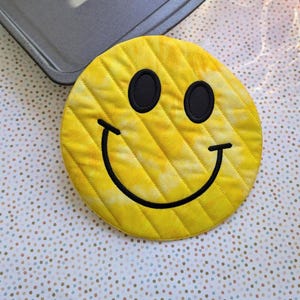 Może przedstawiać: Żółta, pikowana podkładka pod gorące z wyszytą czarną buźką.