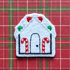 May include: White felt gingerbread house applique with red and green candy details. The house has a black door and two candy canes on either side.