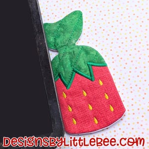 Op de afbeelding: Een rode en groene vilten tas in de vorm van een aardbei met gele stiksels. De tas ligt op een witte achtergrond met roze en oranje stippen.