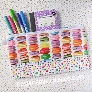 May include: A white zippered pouch with a colourful macaron pattern. The pouch has a white polka dot lining and is filled with colourful pens and a purple mini composition book that says "Mini Composition Book" and "50 Sheets".