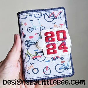 May include: A white fabric notebook cover with a blue denim border. The cover features a repeating pattern of colorful bicycles. A white patch with red embroidery reads "2024 Class of 24".