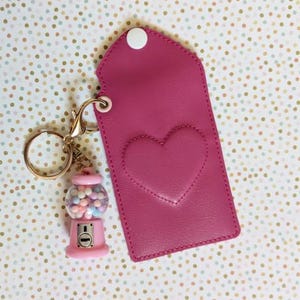 May include: A magenta rectangular faux leather pouch with a white snap closure and a stitched heart design on the front. Attached to the gold-toned keyring is a miniature pink gumball machine charm filled with tiny multi-colored spheres.