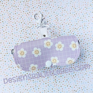 Könnte beinhalten: Eine violette und weiße Reißverschlusstasche mit einem Blumenmuster und Smiley-Gesichtern. Die Tasche hat eine weiße Metallschaufe für die Befestigung eines Schlüsselanhängers.