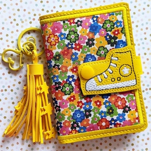 Peut inclure: Un portefeuille en cuir jaune avec un motif floral et une chaussure en cuir jaune avec des coutures blanches et une étoile blanche sur le côté. Le portefeuille a un porte-clés en cuir jaune avec un pompon et un motif d'étoile.