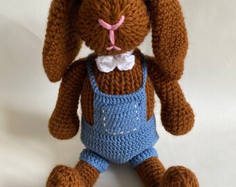 May include: A brown crocheted bunny wearing blue overalls with a white bow tie. The bunny is sitting with its legs crossed.