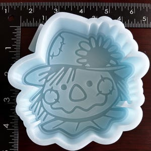以下が含まれることがあります： わら帽子をかぶったかかしの明るい青色のシリコンモールド。ボタンの鼻と優しい笑顔をしています。このモールドは、クラフトやレジンを使ったクラフト制作に最適です。