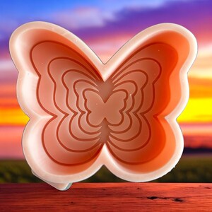 May include: A pink silicone mold shaped like a butterfly with multiple layers. The mold is designed for making soap, candles, or resin crafts.