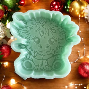 May include: A light green silicone mold in the shape of a Highland cow. The mold has a scalloped edge and features a detailed illustration of the cow with large eyes and a floral crown. Christmas ornaments and lights are in the background.