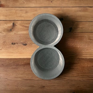 May include: A gray, double-bowl serving dish with a textured interior, resting on a wooden surface. The dish has a figure-eight shape, with two circular bowls connected. The wood grain is visible.