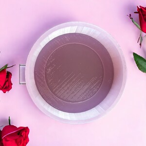 May include: A white cake pan with a textured surface surrounded by red roses on a pink background.