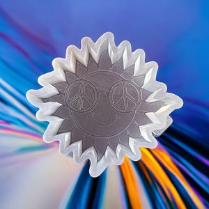 May include: A white sticker with a gray sun design. The sun is wearing peace sign sunglasses and has a smiling face.