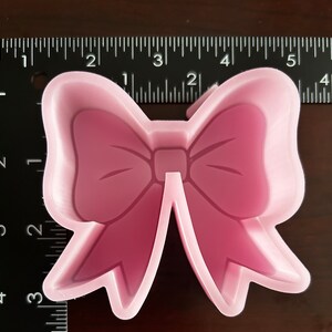 May include: Pink plastic cookie cutter shaped like a bow. The bow is about 2 inches wide.
